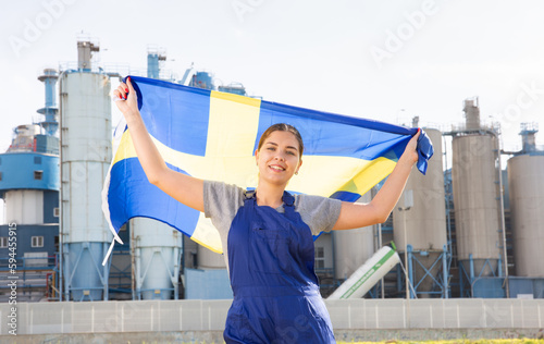 Cheerful young woman in workwear holds the national flag of Sweden in her hands, standing on the street against the background .of an industrial enterprise © JackF
