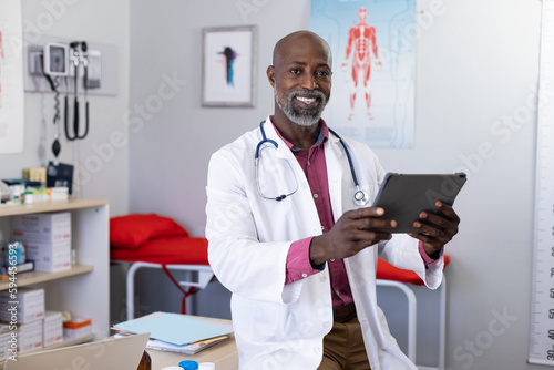 Portrait of happy african american male doctor wearing stethoscope using tablet at doctor's office