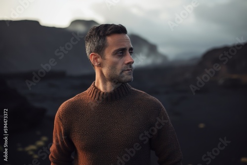 Portrait of a handsome young man with dark hair, wearing a brown sweater, standing in the middle of a volcanic landscape.