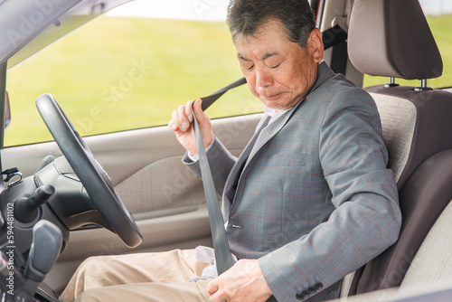 自動車の運転席に乗りシートベルトを締めるアジア人の高齢者ドライバー  © buritora