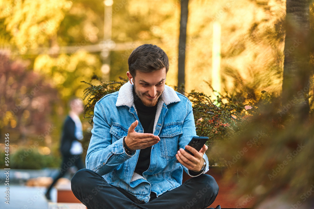 Obraz premium A man sitting on a bench in the city center using his smartphone and wireless headphones for a call, showcasing the convenience of technology in modern communication and the ability to multitask while