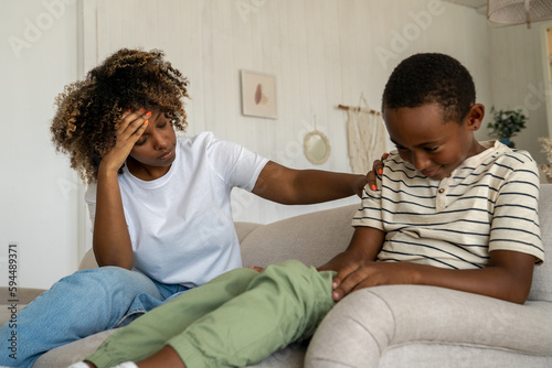Exhausted African American mother grabs head from antics of kid boy who smiles maliciously. Naughty mischievous mixed race child son indulges while sit on couch unnerving ignore upbringing tired woman