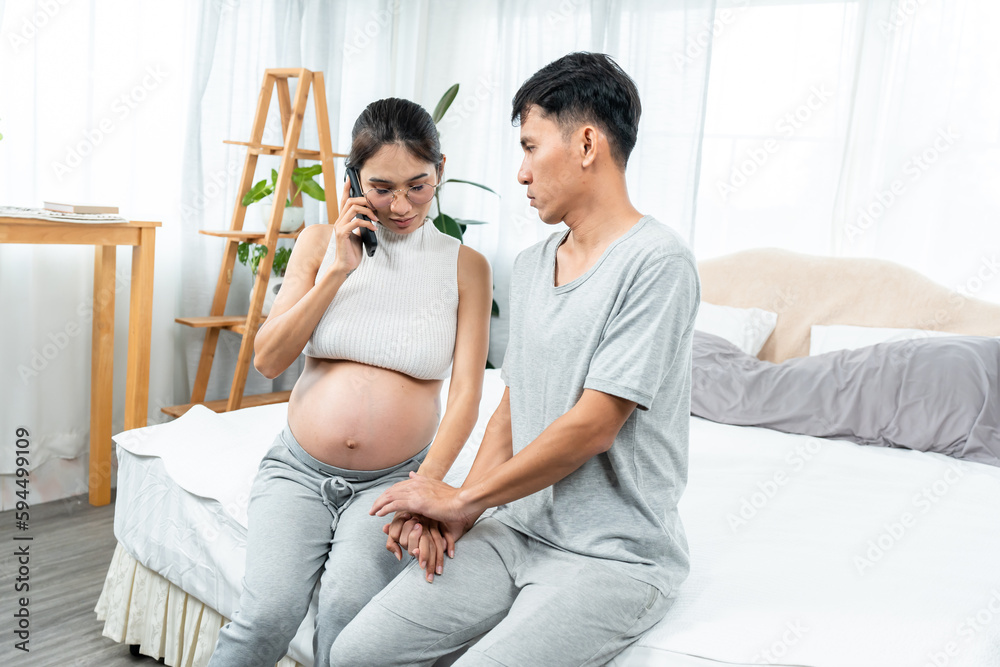 Asian pregnant woman talking on phone with doctor Counseling about pregnancy and there is stress. Husband holding hands and gently stroking wife's hand to relieve stress, comfort wife encourage each Asian pregnant woman talking on phone with doctor Counseling about pregnancy and there is stress. Husband holding hands and gently stroking wife's hand to relieve stress, comfort wife encourage each