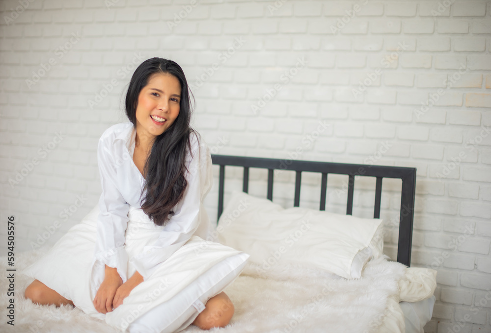 Black haired woman sitting of a modern hotel room in the morning on the bed with both hands holding the pillow and smile at the camera. Black haired woman sitting of a modern hotel room in the morning on the bed with both hands holding the pillow and smile at the camera.