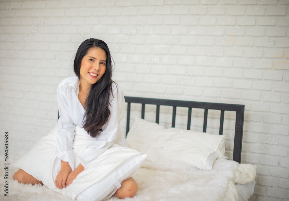 Black haired woman sitting of a luxury modern hotel room in the morning on the bed with both hands holding the pillow and smiles at the camera. Black haired woman sitting of a luxury modern hotel room in the morning on the bed with both hands holding the pillow and smiles at the camera.