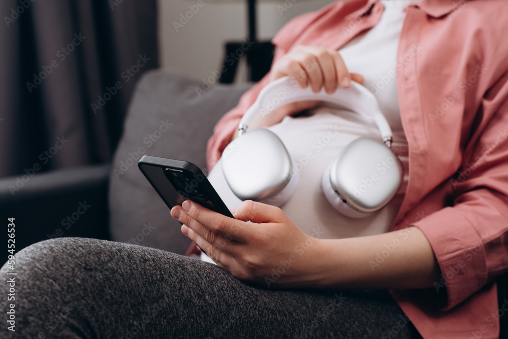 Fototapeta premium Close up of young pregnant woman applying headphones on belly sitting alone on comfy sofa at home. Expectant mother listening music in cozy living room. Future mom giving unborn listening music