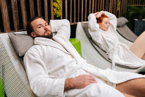 Portrait of young couple in white bathrobes lying on comfortable deck chair by poolside after spa treatment at beauty salon. Brutal man and cute woman enjoying relaxing after body massage treatment.