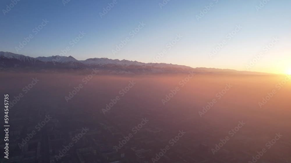 Epic gray smog is visible at sunset over the city. A bird's-eye view from a drone of houses, roads, cars and parks. White clouds and snowy mountains are illuminated by orange rays of the sun. Almaty Epic gray smog is visible at sunset over the city. A bird's-eye view from a drone of houses, roads, cars and parks. White clouds and snowy mountains are illuminated by orange rays of the sun. Almaty