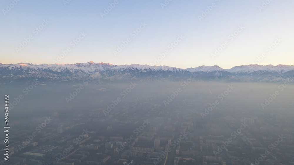 Epic gray smog is visible at sunset over the city. A bird's-eye view from a drone of houses, roads, cars and parks. White clouds and snowy mountains are illuminated by orange rays of the sun. Almaty