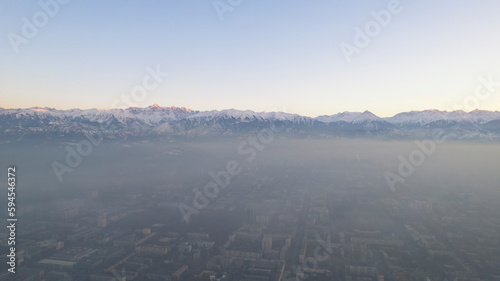 Epic gray smog is visible at sunset over the city. A bird s-eye view from a drone of houses  roads  cars and parks. White clouds and snowy mountains are illuminated by orange rays of the sun. Almaty