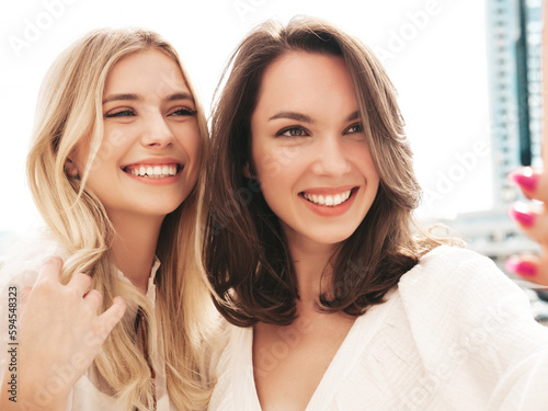 Two young beautiful smiling hipster female in trendy summer white dress clothes. Sexy carefree women walking in street. Positive models having fun, hugging and laughing. Taking selfie photos