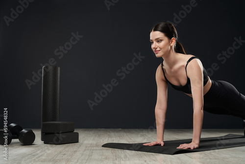 Side view of focused girl working out, training indoors in studio. Attracrtive, sporty, young woman standing in plank on yoga mat, smiling. Isolated on black studio background.