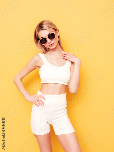 Young beautiful smiling blond female in summer cycling shorts clothes. Sexy carefree woman posing near yellow wall in studio. Positive model having fun indoors. Cheerful and happy. In sunglasses