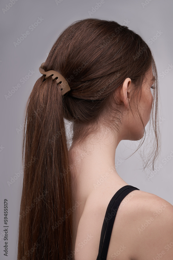 Fototapeta premium A half-turned brown-haired girl with a ponytail is wearing a black tank top. The lady's hair is fixed with a brown chocolate matte banana hair clip. The photo was taken on a light background.