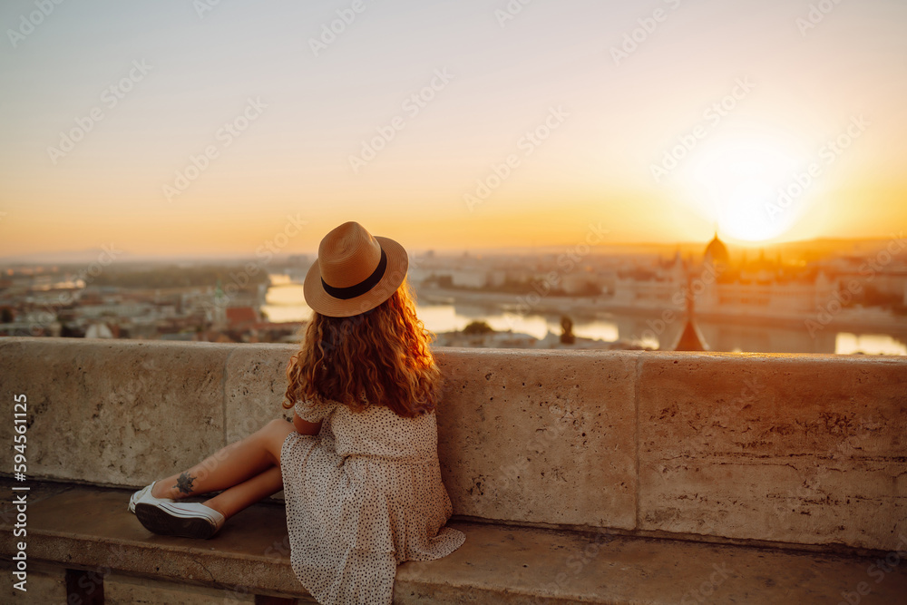 Naklejka premium Smiling female tourist in a hat admires the landscape of the city at dawn. Euro-trip. Travel, tourism and active lifestyle concept.