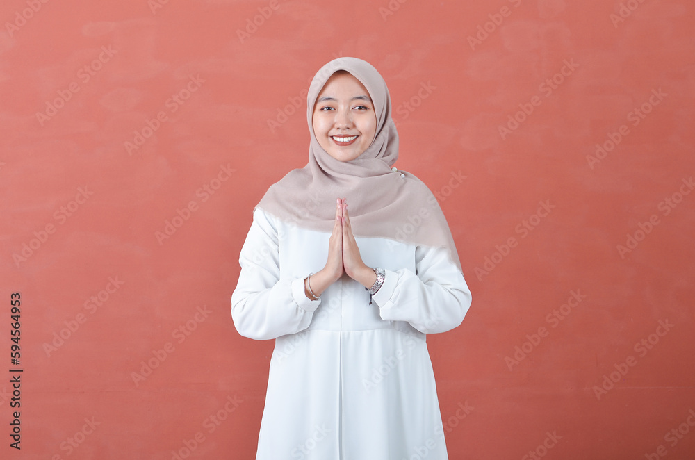Beautiful Asian woman in white gamis and hijab smiling cheerfully showing namaste gesture isolated in brown background Beautiful Asian woman in white gamis and hijab smiling cheerfully showing namaste gesture isolated in brown background