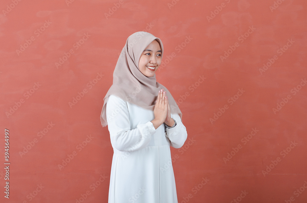 Beautiful Asian woman in white gamis and hijab smiling cheerfully showing namaste gesture isolated in brown background Beautiful Asian woman in white gamis and hijab smiling cheerfully showing namaste gesture isolated in brown background