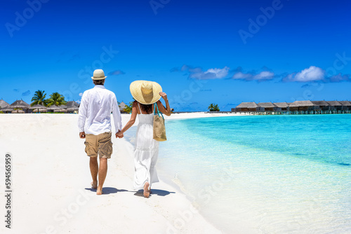 Happy couple in white summer clothing and with hats walks on a tropical paradise beach with turquoise sea in the Maldives islands