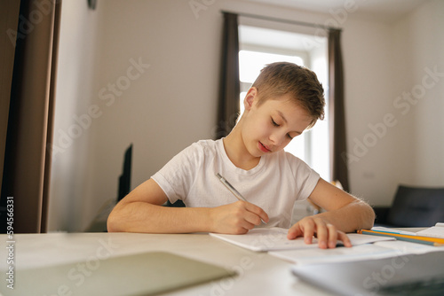 Little kid boy making school homework on laptop and making notes while sitting at home in his room