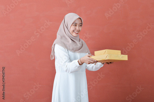 Beautiful Asian woman in white gamis and hijab smiling cheerfully bring a giift box isolated in brown background photo