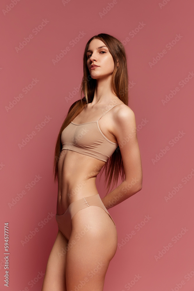 Portrait of young girl with slim body, belly, butticks posing in beige underwear against pink studio background. Concept of beauty, body care, fitness, sport, health, figure, wellness Portrait of young girl with slim body, belly, butticks posing in beige underwear against pink studio background. Concept of beauty, body care, fitness, sport, health, figure, wellness