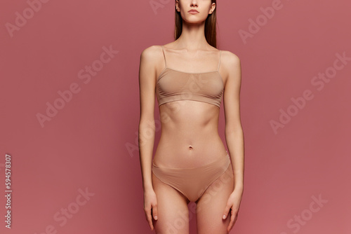 Cropped image of slim, muscular, beautiful female body. Young model posing beige underwear against pink studio background. Concept of beauty, body care, fitness, sport, health, figure, diet