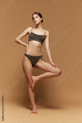 Full-length portrait of young beautiful girl with slim, fit body shape, posing in underwear against light brown studio background. Concept of beauty, body care, fitness, sport, health, figure