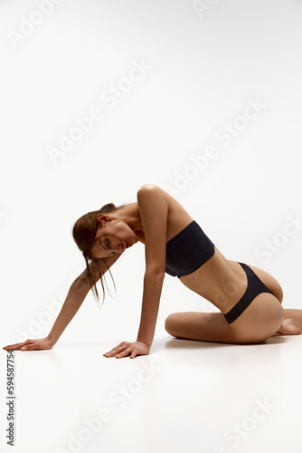 Femininity and well-being. Young girl with fit, slim, healthy body in black underwear posing on floor against white studio background. Concept of beauty, body care, fitness, sport, health, figure
