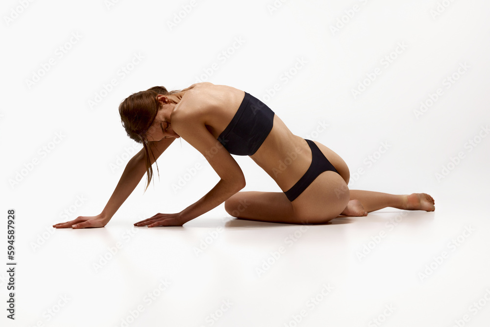 Young girl with fit, slim, healthy body in black underwear posing on floor against white studio background. Taking care after self. Concept of beauty, spa, body care, fitness, sport, health, figure