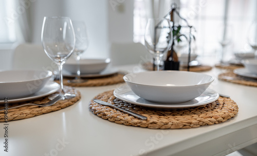 A white set dining room table with white plates  cutlery and glasses on woven place mats. There is a small bonsai in the background.