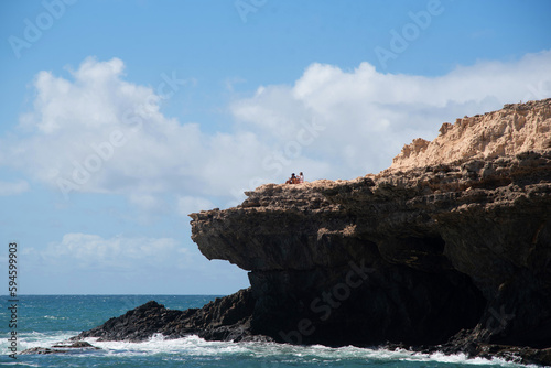 Pareja de turistas sentada en el acantilado de roca volcánica sobre las cuevas de Ajuy disfrutando del día soleado con el tranquilo mar turquesa en Fuerteventura, Islas Canarias. photo