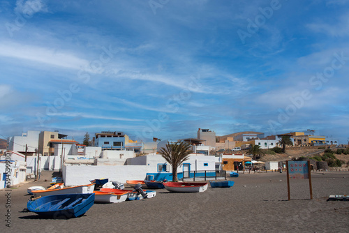 Panorámica de Ajuy en Fuerteventura con coloridos barcos de pesca en la arena negra junto a una palmera y en la parte trasera la arquitectura tradicional de las Islas Canarias