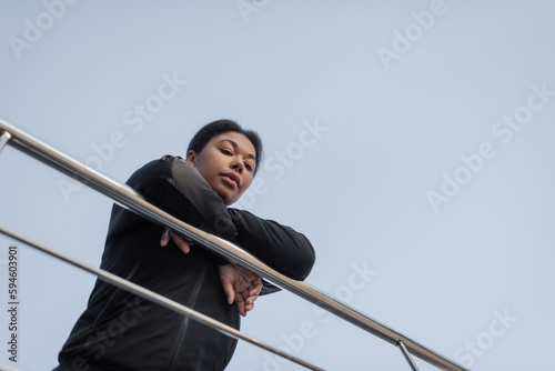 Bottom view of multiracial woman with depression looking at camera near railing on urban street.