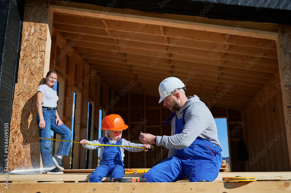 Father, mother and son building wooden frame house. Toddler boy helping his daddy, while woman looking for them on construction site. Guys wearing helmet and blue overalls. Carpentry, family concept. Father, mother and son building wooden frame house. Toddler boy helping his daddy, while woman looking for them on construction site. Guys wearing helmet and blue overalls. Carpentry, family concept.