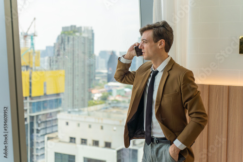Handsome young white businessman talking on the phone and looking out the window view of city skyline in a business district area