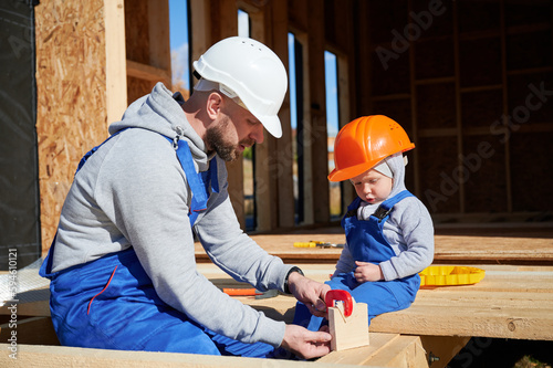 Father with toddler son building wooden frame house. Male builder learing kid to use clamp on construction site, wearing helmet and blue overalls on sunny day. Carpentry and family concept.