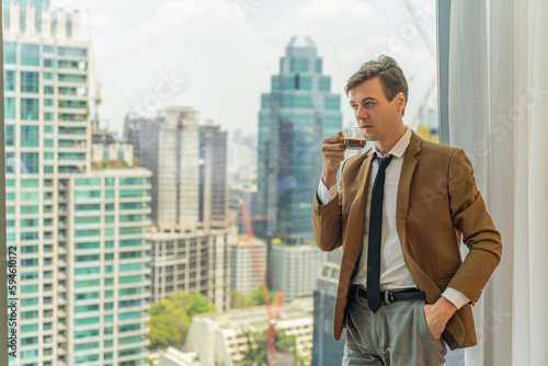 Handsome young white businessman drinking a cup of coffee standing looking out the window view of city skyline in a business district area © asean studio