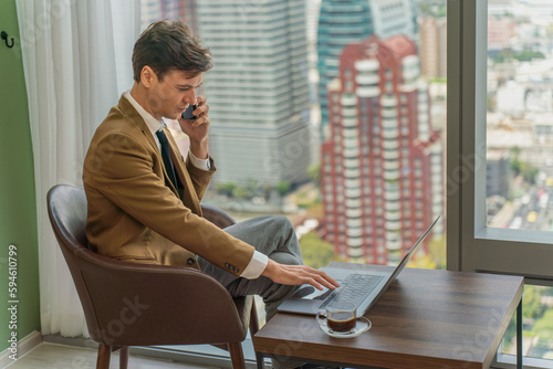 Handsome young white businessman talking on the phone while working on his laptop in front of the window view of city skyline in a business district area