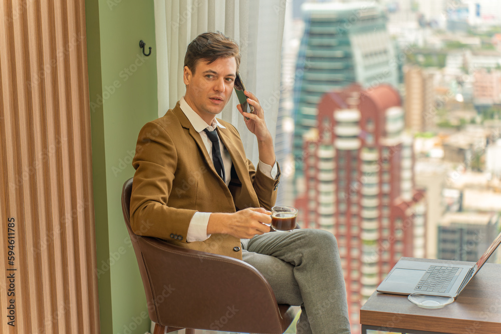 Handsome young white businessman talking on the phone and holding a cup of coffee in front of the window view of city skyline in a business district area Handsome young white businessman talking on the phone and holding a cup of coffee in front of the window view of city skyline in a business district area