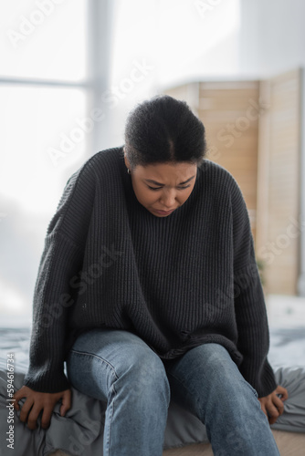 Young multiracial woman with mental problem sitting on bed at home.