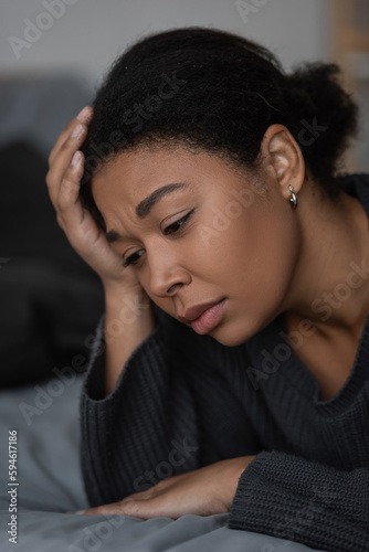 Portrait of upset multiracial woman lying on blurred bed at home.