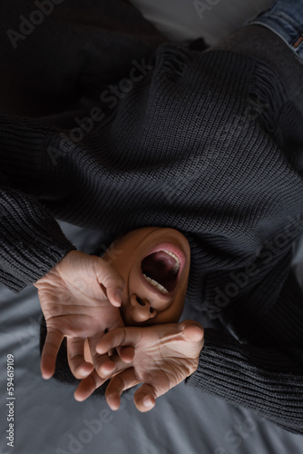 Top view of stressed multiracial woman covering face and screaming on bed at home.