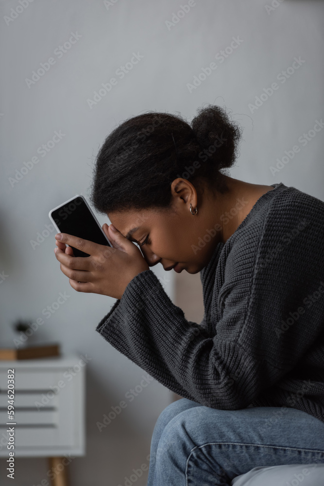 Side view of young multiracial woman with mental problem holding cellphone while sitting on bed at home. Side view of young multiracial woman with mental problem holding cellphone while sitting on bed at home.