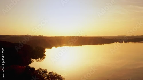 A drone flies backwards over a lake as the sun rises. The landscape is reflected in the water. Location: Germany