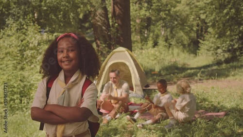 Medium slowmo portrait of smiling African American girl scout in uniform posing for camera with hands folded standing on sunny forest glade, camping with troop and leader in summer