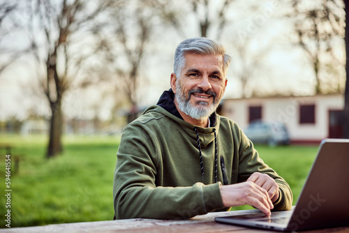 Portrait of a smiling businessman using a laptop while sitting on a wooden bench in the park.