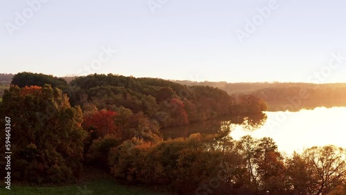 A drone flies towards a forest, a lake stretches out beside it. The trees are reflected in the water and their leaves are colourful. Location: Germany, Season: Autumn