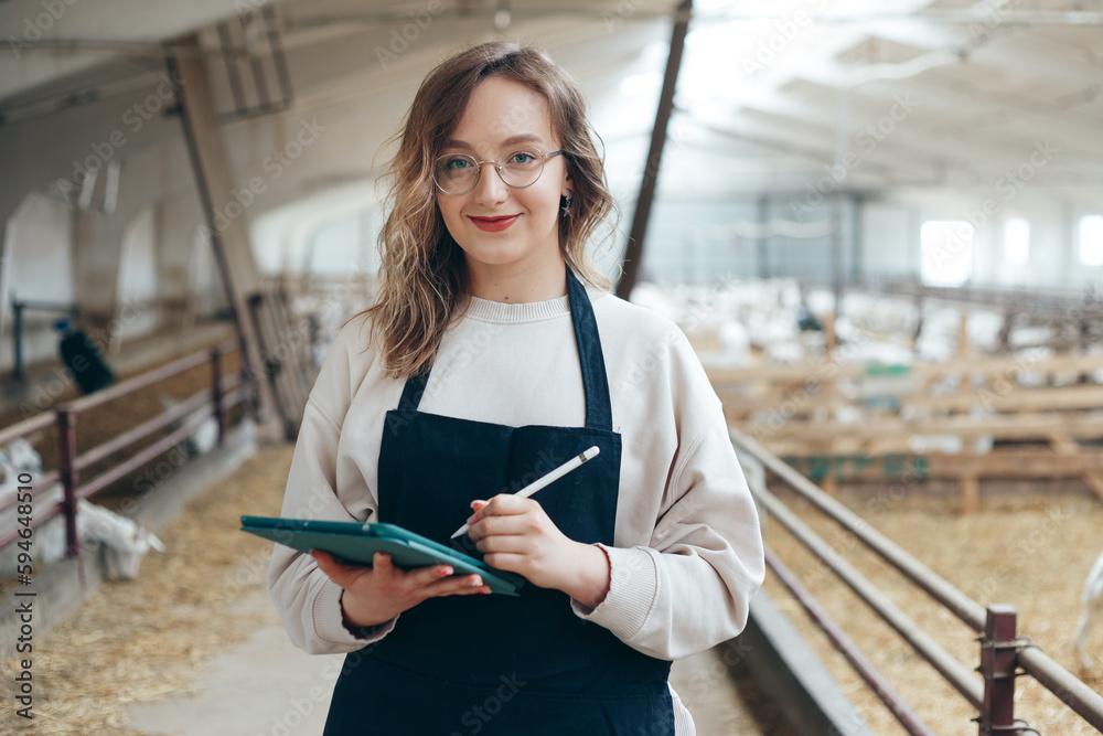 Young Caucasian Female Farm Owner with Tablet in large Livestock Stall with white Dairy Goats