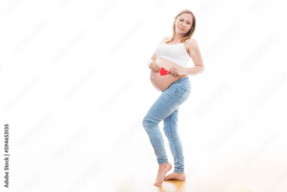 Portret nierozpoznawalnej kobiety podczas ostatnich miesięcy ciąży, trzymającej swój duży brzuch i czerwone serduszko delikatnie stojącej przy ścianie na białym tle - obrazy, fototapety, plakaty 