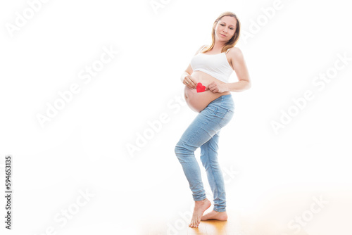 Portret nierozpoznawalnej kobiety podczas ostatnich miesięcy ciąży, trzymającej swój duży brzuch i czerwone serduszko delikatnie stojącej przy ścianie na białym tle